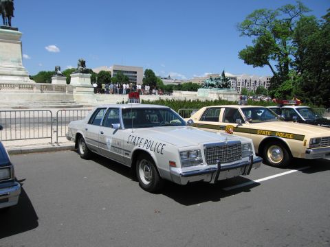 1991 Ford LTD Crown Victoria - silver State Police sedan with push bumper parked near a stone monument, flanked by tan and gold patrol cars.
