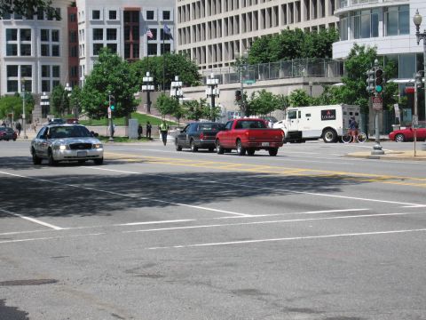 1991 Ford LTD Crown Victoria - silver sedan leads a dark SUV and red pickup down a wide city street past a white Loomis armored truck and office buildings.