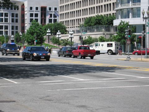 1991 Ford LTD Crown Victoria - Black sedans, a red pickup and a white armored truck move through a wide intersection lined with office buildings and flags.