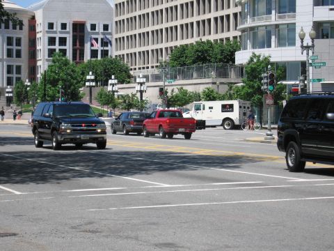 1991 Ford LTD Crown Victoria - Black SUVs and a red pickup move through a downtown intersection near a white armored truck and modern buildings.