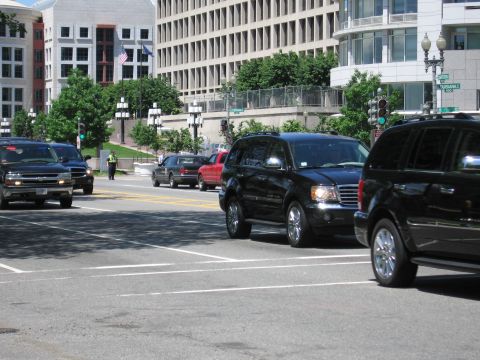1991 Ford LTD Crown Victoria - Two black SUVs turn through a wide city intersection near a traffic-directing officer and modern office buildings.