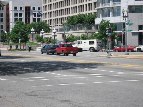 1991 Ford LTD Crown Victoria - dark car and a red pickup cross a wide intersection near a white Loomis armored truck and glass office buildings.