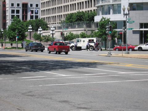 1991 Ford LTD Crown Victoria - dark sedan and red pickup cross a broad city intersection past a parked white armored truck and office buildings.
