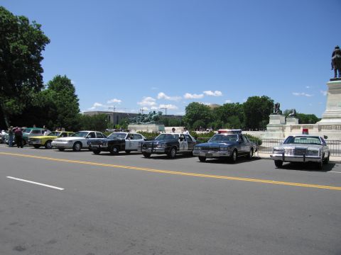 1991 Ford LTD Crown Victoria - row of vintage police cruisers in varied liveries parked along a street in front of a bronze monument and trees.