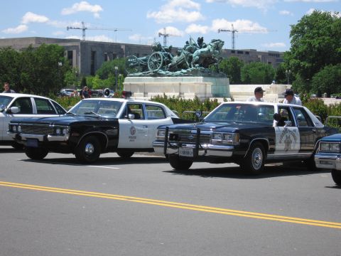 1991 Ford LTD Crown Victoria - Black-and-white police cars, including an LAPD cruiser, parked near a bronze charioteer monument with construction cranes behind.