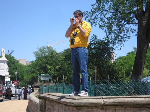 1991 Ford LTD Crown Victoria - man in a yellow hooded sweatshirt and jeans stands on a stone ledge taking a photo, with trees and buildings behind him.