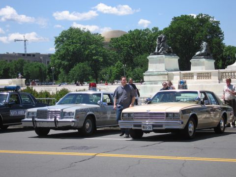 1991 Ford LTD Crown Victoria - man poses between a silver State Police sedan and a gold patrol car parked near a stone monument and lion sculptures.