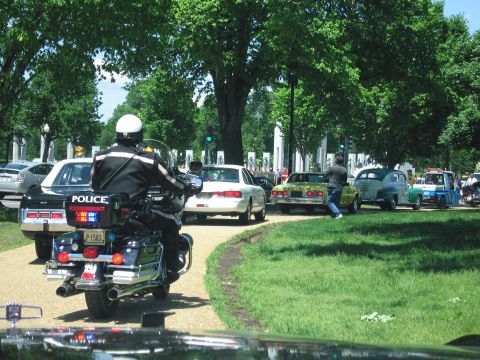 1991 Ford LTD Crown Victoria - uniformed officer rides a police motorcycle along a tree-lined park road behind a line of patrol cars.