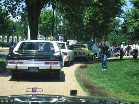 1991 Ford LTD Crown Victoria - rear of a black police car with light bar leads a line of cruisers along a park path as a man photographs them.