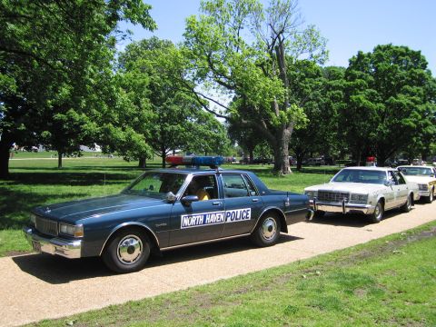 1991 Ford LTD Crown Victoria - blue North Haven Police sedan with light bar parked on a paved park path ahead of other vintage cruisers and trees.