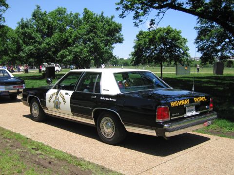1991 Ford LTD Crown Victoria - black-and-white Highway Patrol sedan marked Safety and Service parked on a park path with green trees behind.