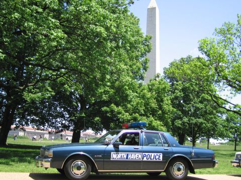 1991 Ford LTD Crown Victoria - blue North Haven Police sedan parked on a park path framed by leafy trees with the Washington Monument behind.