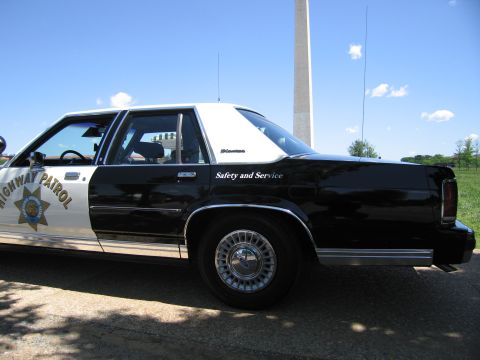 1991 Ford LTD Crown Victoria - Close side view of a black-and-white Highway Patrol sedan reading "Safety and Service," with a monument and blue sky behind.