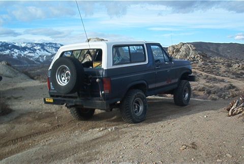 1986 Ford Bronco - This 1986 Ford Bronco is shown in a rear three-quarter view, parked on a dirt road with a mountainous landscape in the background.