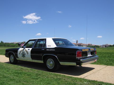 1991 Ford LTD Crown Victoria - Rear-quarter view of a black-and-white Highway Patrol sedan parked on grass with trees and buildings behind.