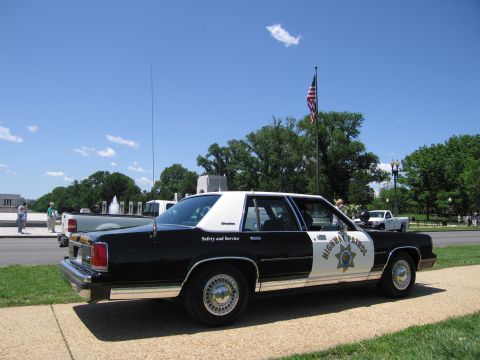 1991 Ford LTD Crown Victoria - black-and-white Highway Patrol sedan parked near a road with a US flag, fountains and trees in the background.
