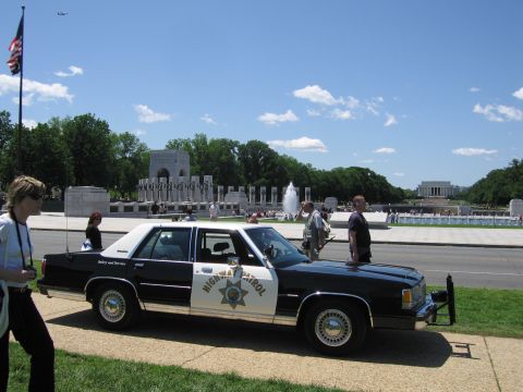 1991 Ford LTD Crown Victoria - black-and-white Highway Patrol sedan with push bar parked near the WWII Memorial as people walk by under a US flag.