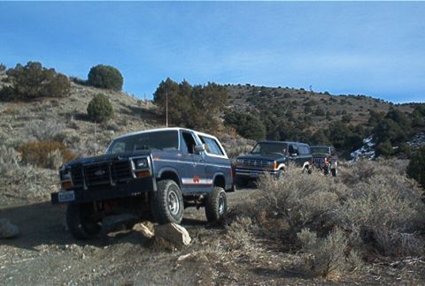 1986 Ford Bronco - Two SUVs navigate a , rocky landscape under a clear blue sky.