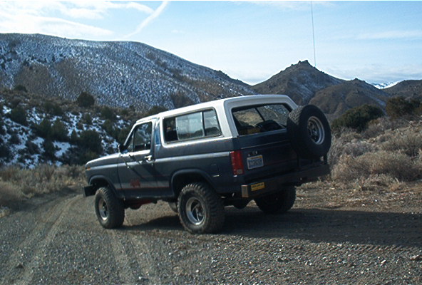 Two-tone 1980s Ford Bronco with rear spare tire carrier on a gravel road below snow-dusted desert hills
