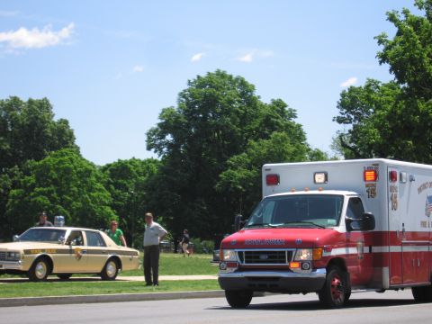 1991 Ford LTD Crown Victoria - red-and-white fire department ambulance drives past while a tan state police sedan and bystanders wait beside a grassy roadside.