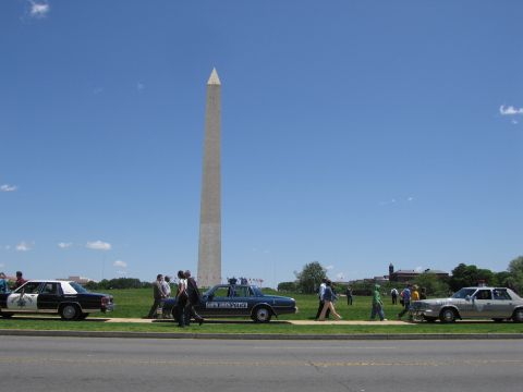 1991 Ford LTD Crown Victoria - Several police sedans and walking pedestrians line a grassy area in front of the towering Washington Monument.