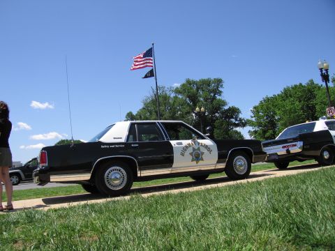 1991 Ford LTD Crown Victoria - black-and-white Highway Patrol sedan is parked along a curb near a flying American flag, with another police car behind.