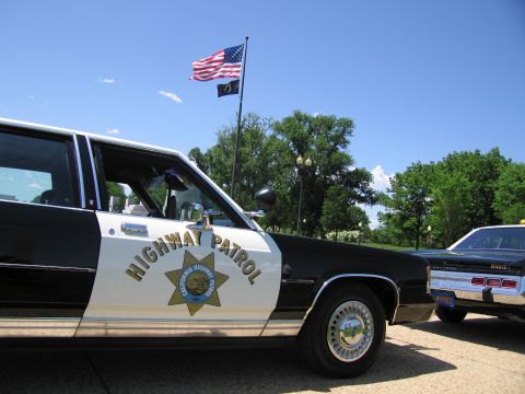 1991 Ford LTD Crown Victoria - black-and-white California Highway Patrol sedan is parked next to a red classic muscle car under an American flag.