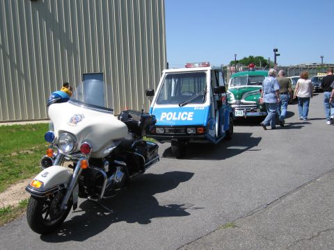 1991 Ford LTD Crown Victoria - white police touring motorcycle beside a small blue three-wheeled Police vehicle parked by a building, with classic cars behind.