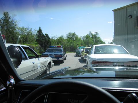 1991 Ford LTD Crown Victoria - Looking out a windshield at a State Police sedan and other vehicles parked along a tree-lined driveway by a building.