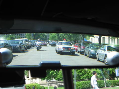 1991 Ford LTD Crown Victoria - Through-the-rear-glass view of a city street showing a police motorcycle and sedans amid rows of parked cars.