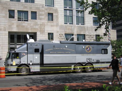 1991 Ford LTD Crown Victoria - large gray FBI Mobile Command Center truck parked behind tape at the curb of a multi-story stone office building.