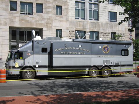 1991 Ford LTD Crown Victoria - gray FBI Mobile Command Center truck with roof satellite dish parked behind yellow tape beside a stone office building.