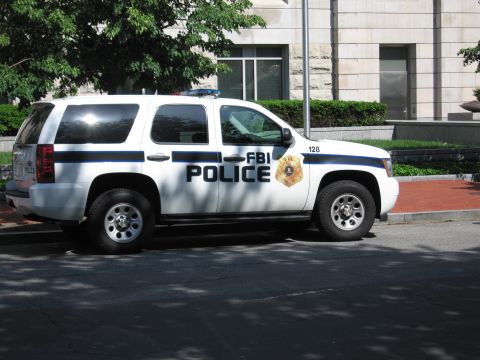 1991 Ford LTD Crown Victoria - white Chevrolet Tahoe SUV lettered FBI Police with a gold badge and unit 128 parked along a curb by a stone building.