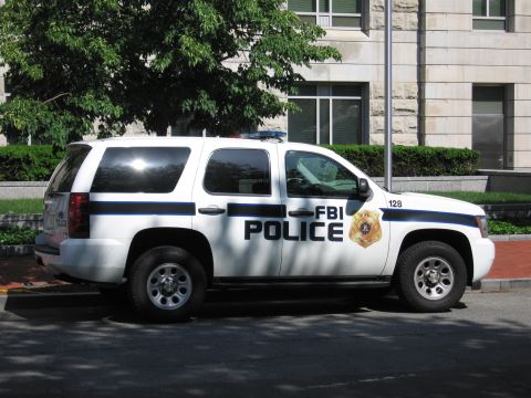 1991 Ford LTD Crown Victoria - Profile of a white Chevrolet Tahoe SUV marked FBI Police with badge and unit 128, parked at a tree-lined curb.