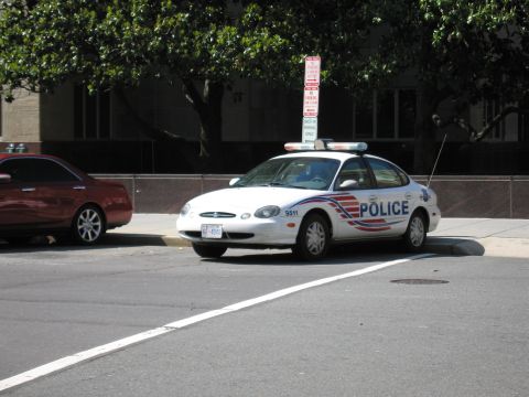 1991 Ford LTD Crown Victoria - white Ford Taurus sedan with a light bar and POLICE markings parked at a curb beside a red sedan.