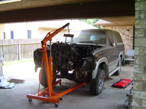 1986 Ford Bronco - A dark gray Ford Bronco is undergoing engine removal in a garage, with an orange engine hoist holding the engine.