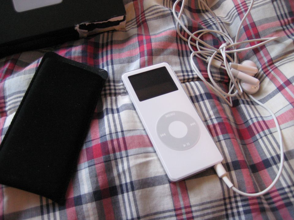 White iPod, black case, and earbuds on a plaid surface. Supermotors.net logo in corner