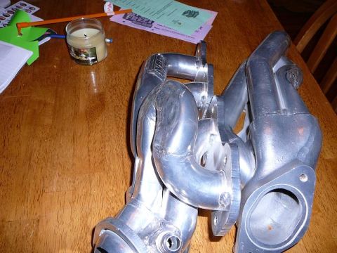 1997 Mercury Grand Marquis - A pair of polished cast aluminum exhaust manifolds/headers sitting on a wooden kitchen table, showing the curved runners, bolt flanges and…