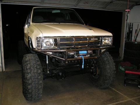 1986 Toyota Hilux - Beige early Toyota 4x4 pickup lifted on mud tires with a tubular front bumper and winch, in a dim garage.