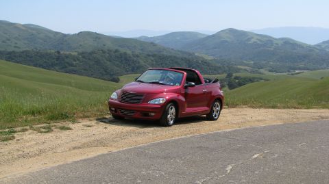 2005 Chrysler PT Cruiser - A red convertible Chrysler PT Cruiser is parked on a dirt road with a scenic backdrop of rolling hills.