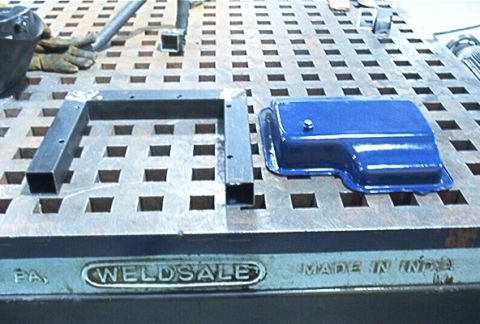 1986 Ford Bronco - A blue transmission pan and a metal frame are placed on a welding table, with 'WELDSALE' visible on the table's edge.