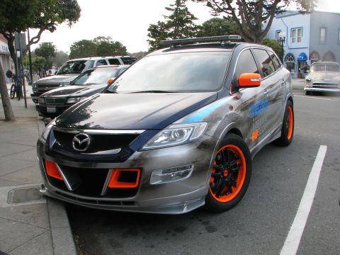 2005 Chrysler PT Cruiser - A modified Mazda CX-9 with bright orange wheels and mirrors is parked curbside.