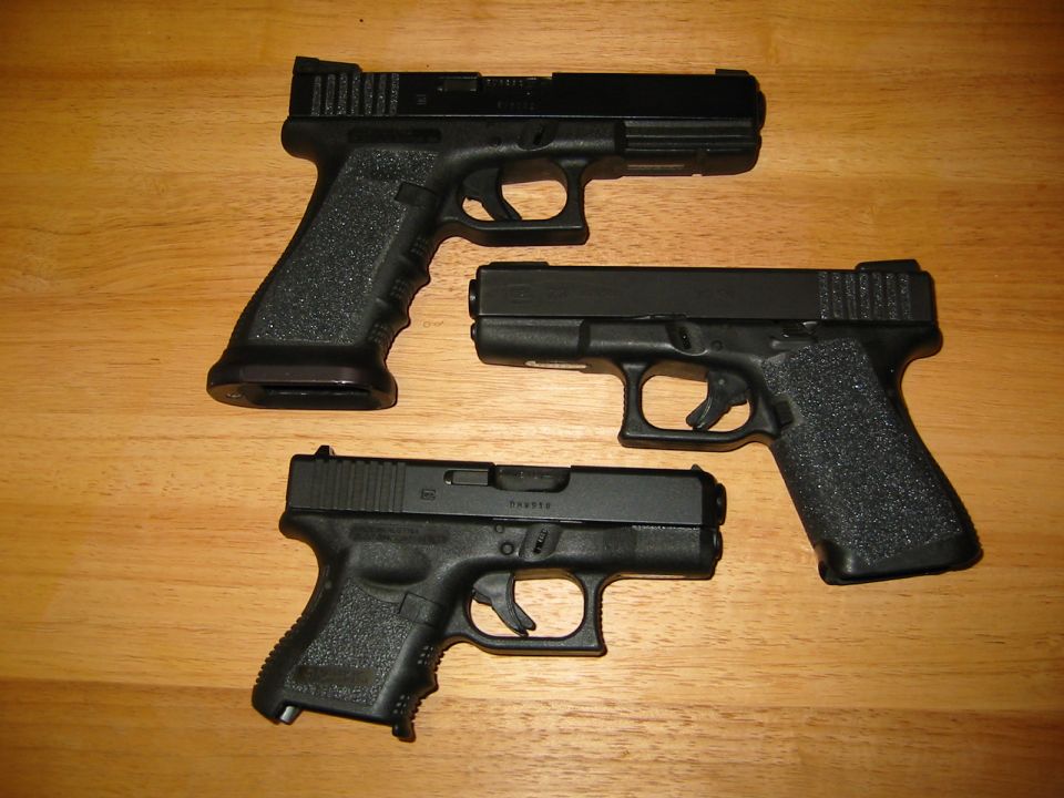 Three black handguns on a wooden surface. The largest handgun is at the top, a medium one is to its right, and a compact one is at the bottom