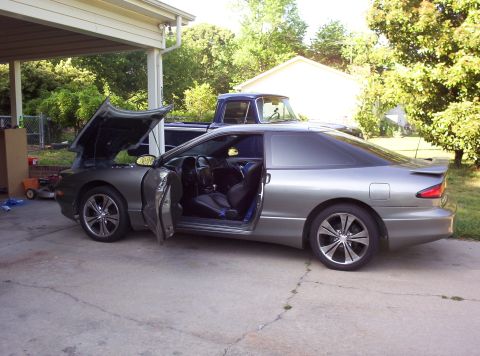 1995 Ford Probe - A gray coupe with its hood and driver's door open is parked in a driveway, with a blue pickup truck behind it.