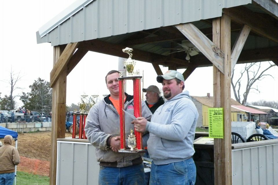 Two men holding a large trophy under a wooden pavilion, with other trophies and vehicles in the background
