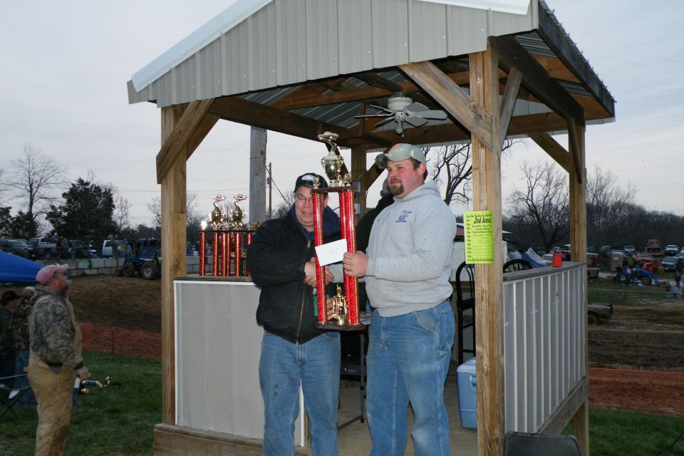Two men holding a large trophy under a covered structure, with other trophies and vehicles in the background