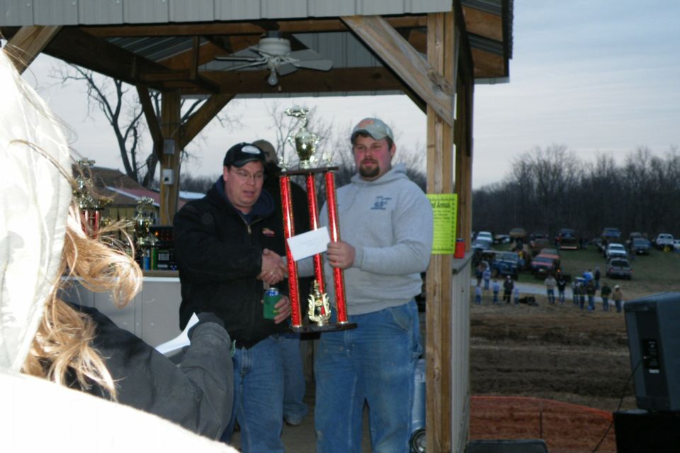 Two men holding a large trophy, one shaking hands, with a crowd and parked vehicles in the background