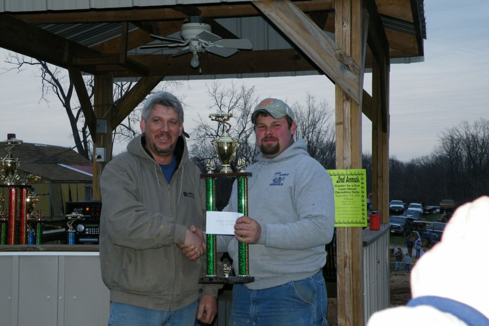Two men shake hands while holding a trophy, with parked cars in the background
