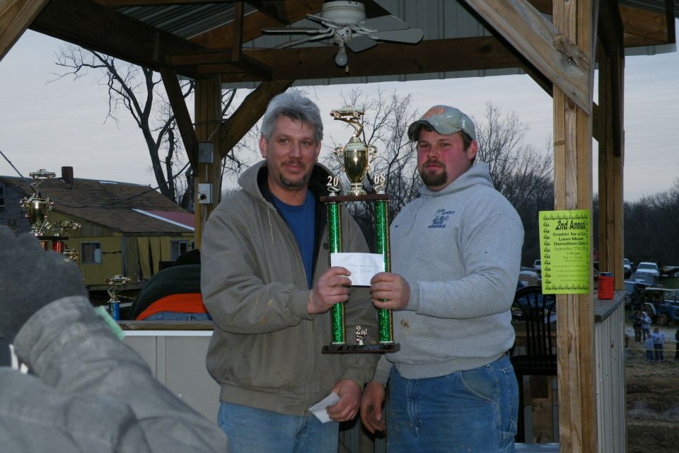 Two men hold a trophy with a check. A yellow sign is on a wooden post