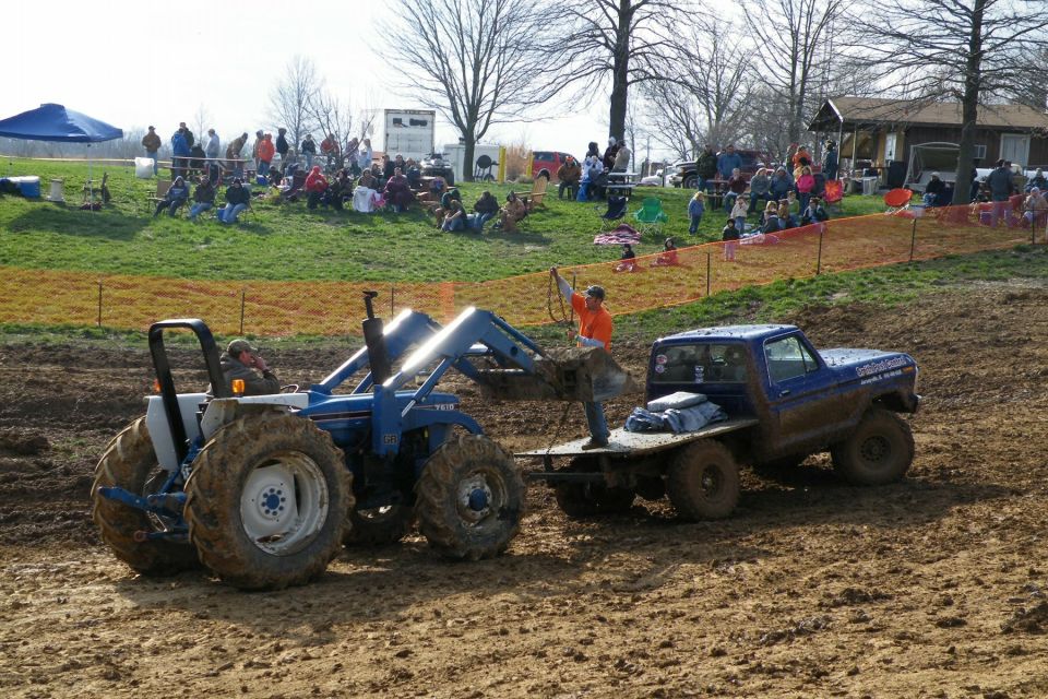 Blue tractor with front loader pulling a blue flatbed truck through mud. Spectators watch from a grassy hill